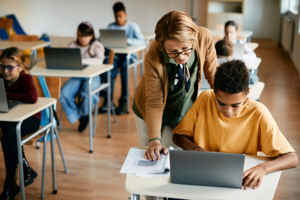 African American schoolboy uses laptop with help of his teacher during computer class in classroom.